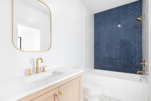 A beautiful renovated bathroom with a natural wood vanity cabinet, gold mirror and faucets, and a blue herringbone tiled shower accent wall.
